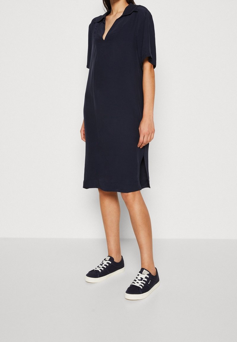 Robe bleu marine à manches courtes avec col polo et fentes sur les côtés, associée à des baskets bleu marine avec des lacets blancs et une semelle blanche.