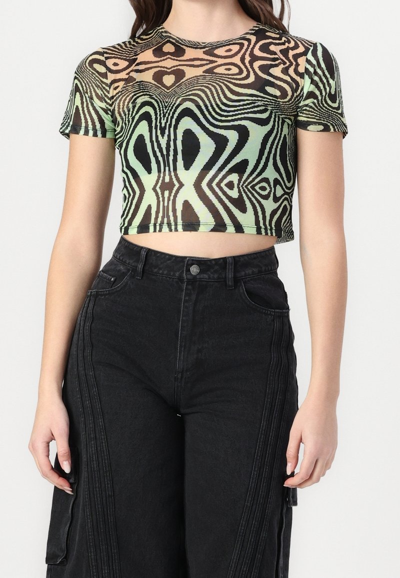 Femme portant un crop top moulant à manches courtes avec un motif abstrait en spirale vert et marron, associé à un pantalon noir taille haute.