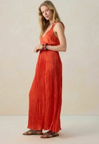 Robe maxi sans manches rouge avec une texture plissée, dotée d'un décolleté en V. Associée à des sandales imprimé léopard et à des bracelets empilés.