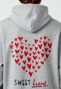 Sudadera gris con un gran diseño bordado de un corazón rojo en la parte posterior, rodeado de corazones más pequeños, con la palabra "DULCE" en escritura negra.