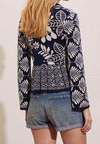 Žena s dlouhými blond vlasy, oblečená v námořnickém modrém svetru s květinovým potiskem a ohrnutých džínových šortkách, zobrazená zezadu.