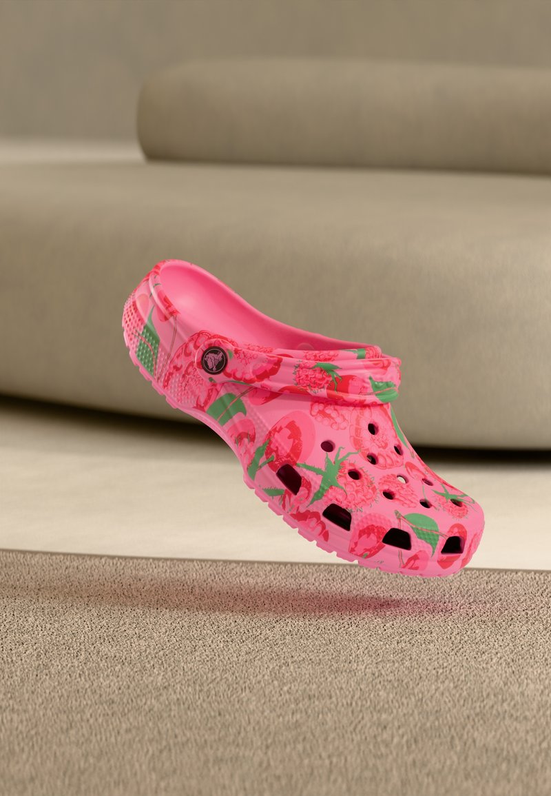 Sabot rose avec motifs de framboises et de feuilles vertes, doté de trous de ventilation et d'une bride arrière, placé à l'intérieur près de meubles beiges.