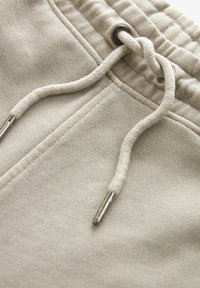 Beige szabadidőnadrág texturált anyagból, fém végződéssel ellátott húzózsinórral és elülső zsebbeli nyílással. Egyszerű és funkcionális dizájn.