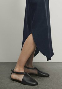 Mustat nahkatossut, joissa on kaareva varvas ja nilkkahihna, yhdessä tummansinisen hameen kanssa, jossa on korkea halkio ja hillitty kiilto.