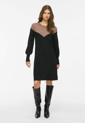 Modelo posa frente a un fondo blanco luciendo un vestido negro hasta la rodilla con canesú marrón en forma de V y botas negras altas.