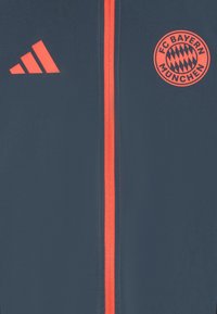 Giacca del FC Bayern München, in tessuto blu navy, chiusura con zip e dettagli in arancione neon, include il logo Adidas e un emblema circolare della squadra.
