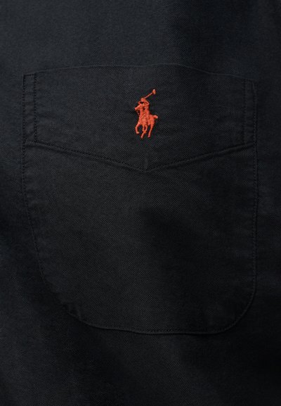 Czarna kieszeń z haftowanym na czerwono logo gracza w polo na środku przedniej części kieszeni, widoczne przeszycia.