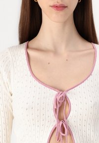 Vitt stickat linne med djup V-hals, rosa kant längs halsringningen och knytning i mitten. Har små diamantmönster.