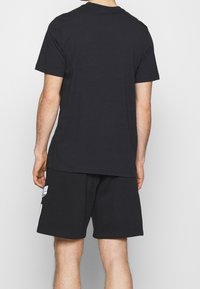 Muž v jednobarevném černém tričku s krátkým rukávem a černých kraťasech s bílým logem na boku, stojící zády k jednoduchému pozadí.