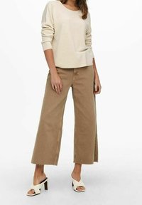 Haut beige à manches longues, pantalon large taille haute tan à coupe ample avec ourlet effiloché, et sandales à talons blanches. Design minimal, lignes simples et textures douces.
