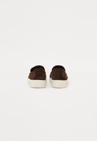 Brune semskede slip-on sko med hvit gummisåle og svarte detaljer. Har en avrundet tå og et minimalistisk design.