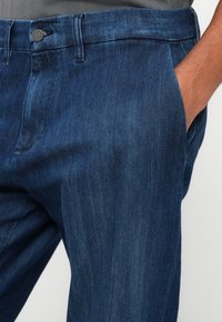 Mørkeblå denimbukser med en glat tekstur, der har en klassisk pasform, en frontlomme og en metalknap i taljen.