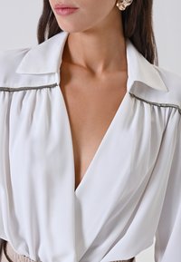 Blusa bianca in seta con scollo a V profondo e colletto strutturato. Presenta tessuto arricciato alle spalle, accentuato da una finitura metallica.