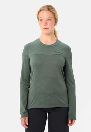 Junge Frau mit lockigem Haar, die ein langärmliges, olivgrünes Vaude-Shirt und schwarze Hosen trägt, stehend vor einem schlichten weißen Hintergrund.