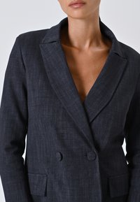 Veste noire en laine à double boutonnage avec revers, deux boutons et poches latérales. Fabriquée dans un tissu texturé et léger.