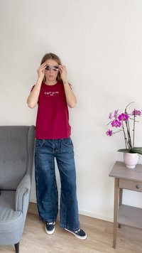 Camiseta burdeos con impresión "C'est la vie Paris", combinada con jeans azules holgados y zapatillas negras; persona ajusta las gafas de sol, junto a una planta en flor.