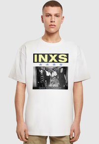 Hvid oversized t-shirt med et sort-hvidt grafisk design af fire personer med "INXS" i gult og "NEED YOU TONIGHT" i sort.