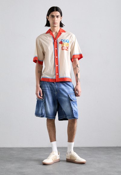 Chemise beige à manches courtes avec des accents rouges et une bordure bleue, comportant un texte coloré. Associée à un short en jean ample et des chaussettes blanches. Baskets beige.