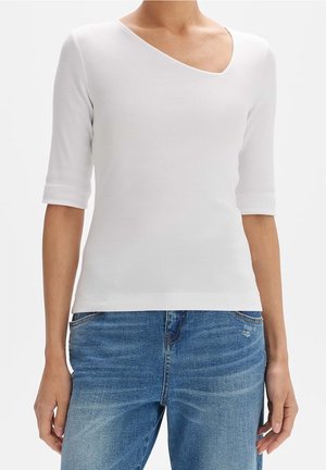 Vrouw draagt een wit geribbeld topje met korte mouwen en een asymmetrische halslijn, gecombineerd met blauw vervaagde jeans.