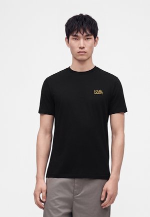 Młody mężczyzna z krótkimi czarnymi włosami, ubrany w czarny t-shirt Karl Lagerfeld i szare spodnie, stoi na jednolitym białym tle.