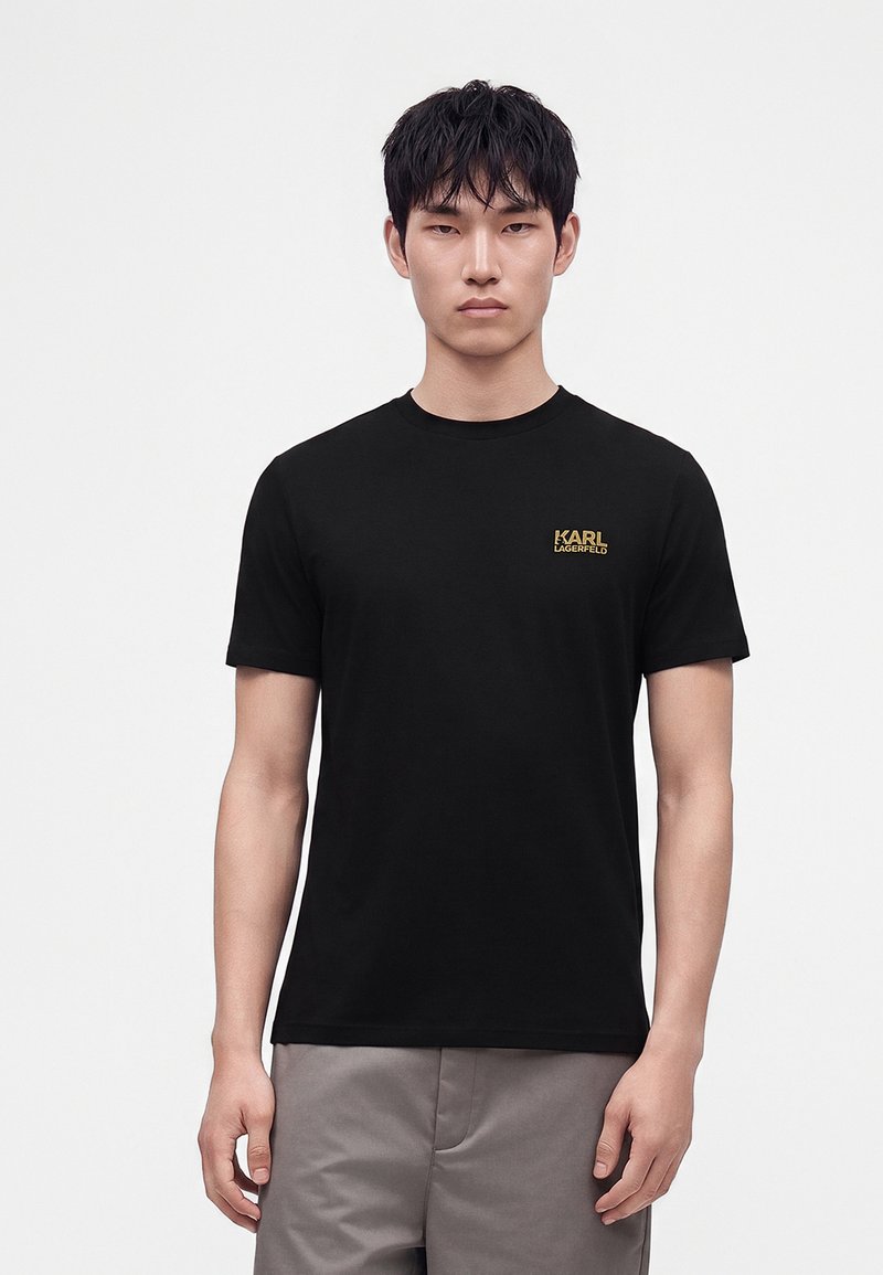 Jeune homme aux cheveux courts noirs portant un t-shirt noir Karl Lagerfeld et un pantalon gris, debout devant un fond blanc uni.