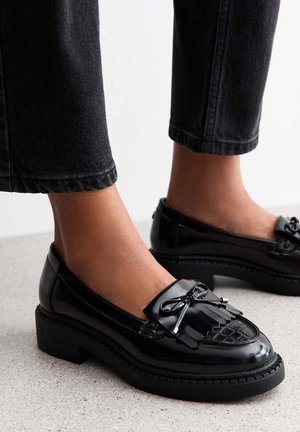 Black patent leather loafers with low block heels, decorative tassels, and fringe worn with cropped black jeans on a light floor.