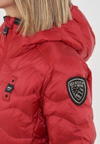 Donna che indossa una giacca trapuntata rossa con cappuccio e una toppa nera con strass sul maniche superiore.
