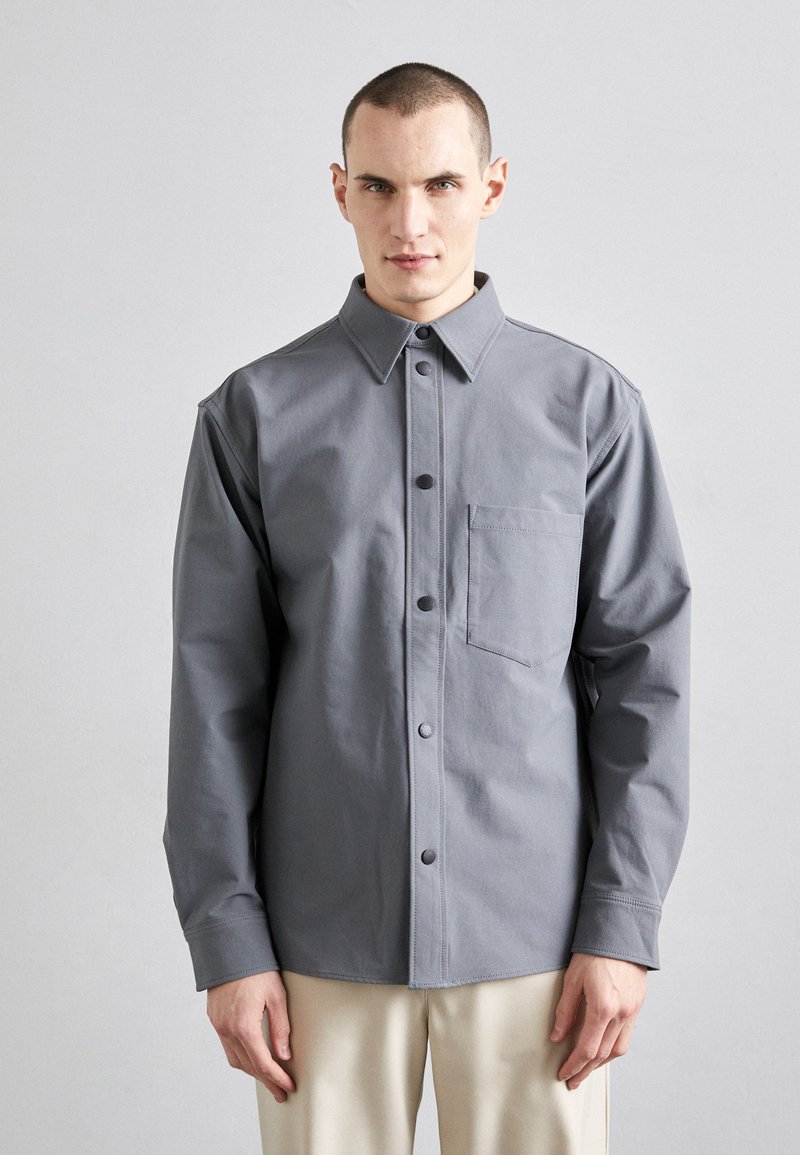 Gray button-up shirt made of fabric with a smooth texture, featuring a pointed collar, front pocket, and matching buttons.