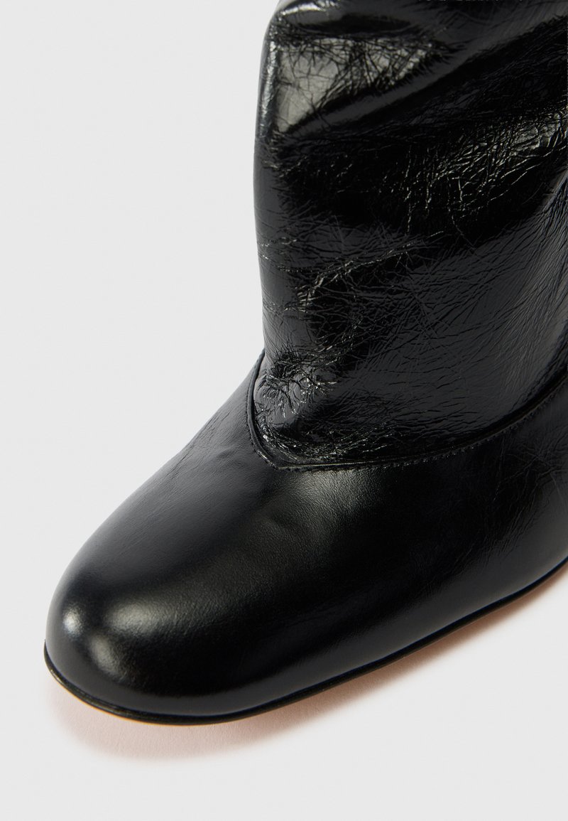 Stivale alla caviglia in pelle nera con finitura liscia e texture leggermente stropicciata, dotato di punta rotonda e dettagli di cucitura minimali.