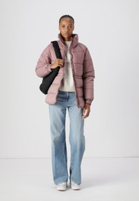 Lys rosa puffjakke med horisontale sømmer, brukt over en kremfarget genser, sammen med blå flagrende jeans og hvite sneakers. Svart veske over skulderen.