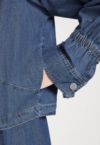 Blå denimjacka med samlad ärmslut, silverknappdetalj och framficka. Slät textur med standard kragsdesign.