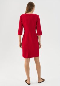 Rotes Kleid mit Dreiviertelärmeln, Reißverschluss hinten, geradem Schnitt und seitlichen Schlitzen. Getragen mit schwarzen Sandalen mit Leopardenmuster.