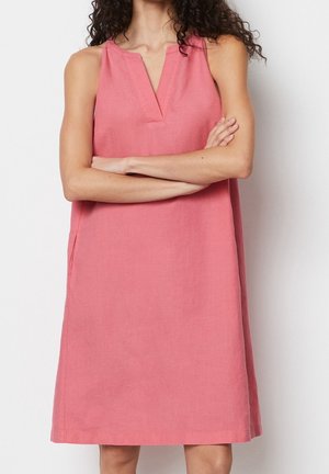 Vrouw met krullend donker haar, gekleed in een mouwloze roze jurk met een V-hals, armen gekruist tegen een witte achtergrond.