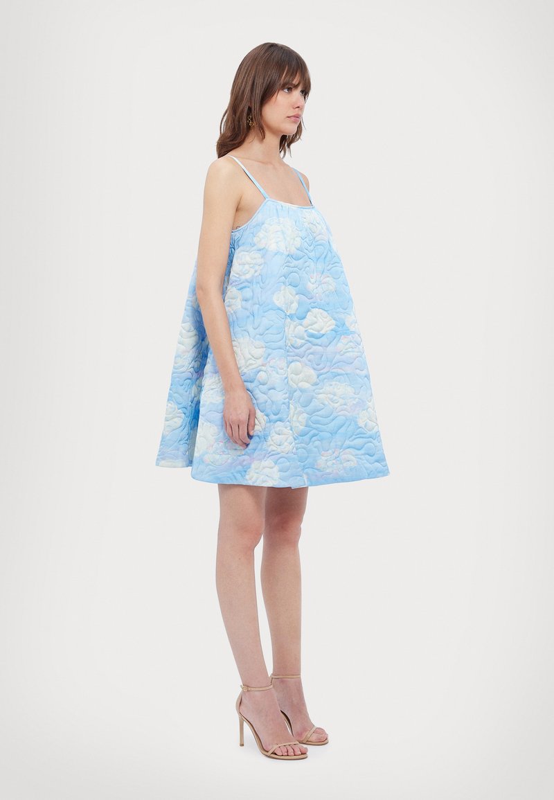 Vrouw staat in een mouwloze, gewatteerde blauwe jurk met wolkenpatroon en dunne bandjes, draagt beige hoge hakken op een effen achtergrond.