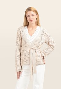 Beige Strickjacke mit offenem Muster, langen Ärmeln und Bindeband in der Taille. Über einem weißen Top mit Spitzenbesatz getragen.