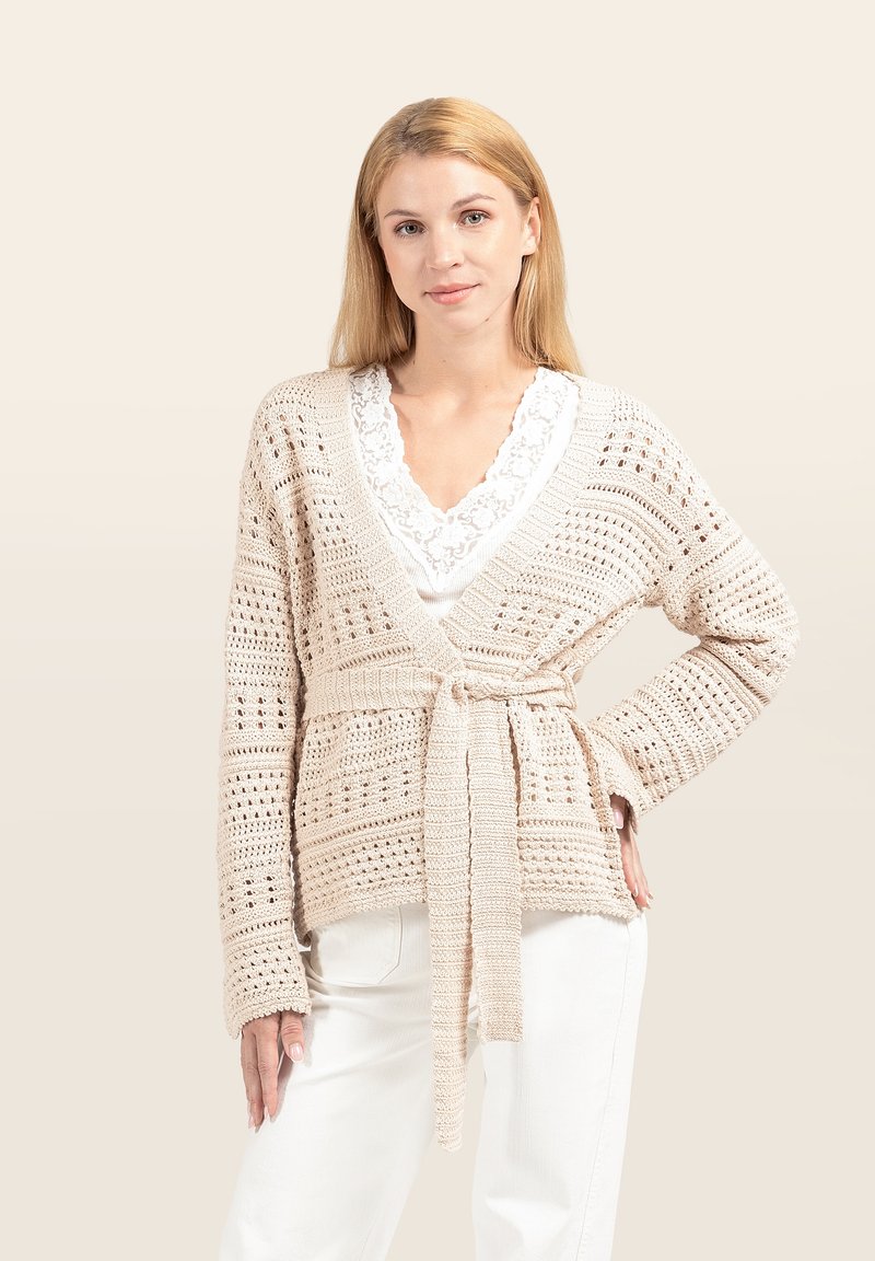 Beige Strickjacke mit offenem Muster, langen Ärmeln und Bindeband in der Taille. Über einem weißen Top mit Spitzenbesatz getragen.