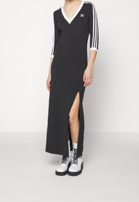 Svart, lång, åtsittande klänning med V-ringning, trekvartsärmar, vita sidostråk och en hög skåra vid låret; bärs med vita snörstövlar.