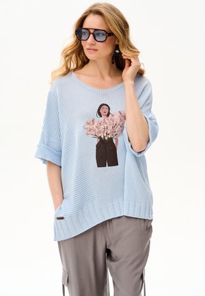 Kobieta o długich blond włosach, nosząca duże okulary w oprawkach w panterkę, jasnoniebieski sweter z wzorem kwiatowym oraz luźne szare spodnie.