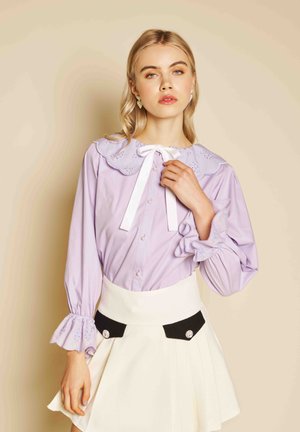 Femme portant un chemisier lavande avec col et poignets à volants, un nœud papillon blanc et une jupe blanche avec des poches boutonnées noires.