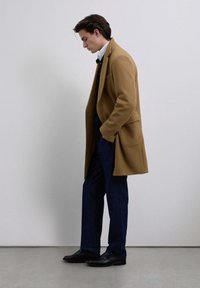 Hombre con abrigo color crema, camisa blanca, suéter oscuro, pantalones azul marino y zapatos negros, de pie con las manos en los bolsillos, mirando hacia abajo contra un fondo liso.