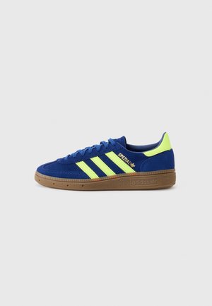 Niebieski sneaker Adidas Spezial z neonowo żółtymi paskami i zakładką na pięcie, brązową teksturowaną podeszwą oraz niebieskimi sznurówkami, pokazany z boku na białym tle.