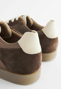 Brune ruskinds sneakers med hvide læderhælelementer og lysebrune snørebånd. Rund tåform og struktureret gummisål.