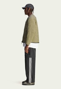 Oliwkowa pikowana kurtka z czarnymi akcentami, nałożona na białą koszulę; zestawiona z ciemnymi spodniami z białymi paskami i czarnymi trampkami.