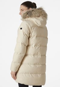 Béžový zateplený puffer kabát s odnímatelnou kapucí s kožešinovým lemováním, dlouhými rukávy a hladkou texturou. Obsahuje boční kapsy a logo na rukávu.