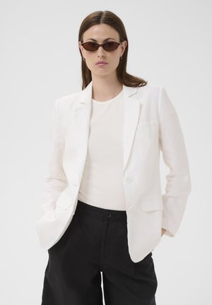 Woman wearing a white blazer, white top, black pants, and slim dark sunglasses, standing with hands in pockets against a plain background.