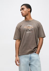 Καφέ κοντομάνικη μπλούζα με γραφικό τύπωμα που απεικονίζει το "LAS VEGAS STRIP" και ένα αυτοκίνητο, συνδυασμένη με ανοιχτόχρωμο τζιν παντελόνι. Μαλακό βαμβακερό υλικό.