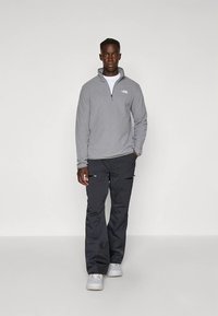 Pullover in pile grigio con zip a quarto e logo sul petto, abbinato a pantaloni neri con tasche con zip e sneaker bianche.