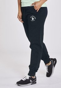 Pantalones de chándal de color navy con logo en el bolsillo izquierdo; cintura elástica y puños. Llevados con zapatos deportivos negros con acentos blancos.