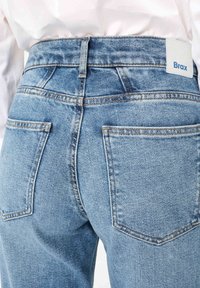 Hellblaue Jeans aus Denim mit zwei Gesäßtaschen und einem weißen Etikett mit der Aufschrift "Brax", getragen zu einem weißen Hemd.