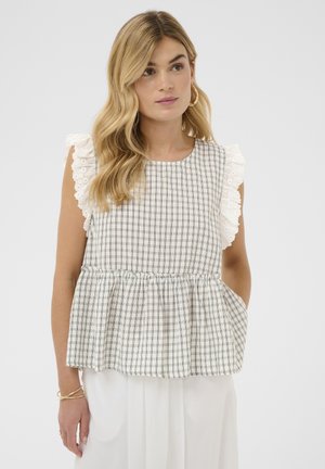 Mujer rubia vestida con blusa sin mangas a cuadros con mangas de encaje blanco con volantes y pantalones blancos, de pie frente a un fondo liso.